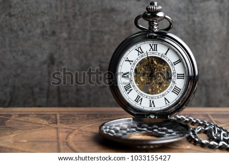 Time symbol or business deadline countdown concept, closed up vintage pocket watch on brown old book with black dark cement wall.