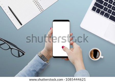 Woman hands holding smartphone with empty screen on blue office desk, surrounded with coffee, laptop, glasses and notebook