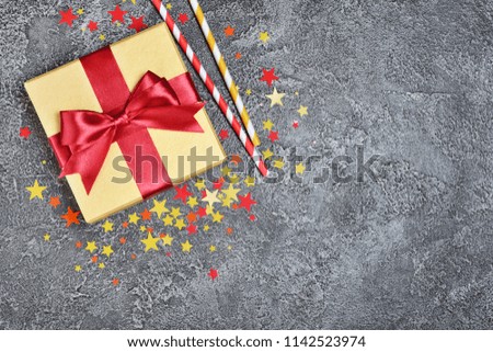 Golden shiny classic gift box with red satin bow and paper cocktail straws with confetti in the shape of stars as attributes of party on gray concrete background