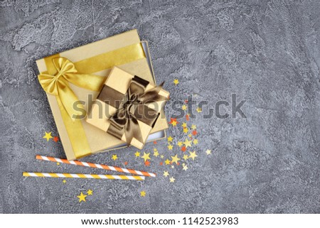 Golden shiny classic gift boxes with brown satin bows and paper cocktail straws with confetti in the shape of stars as attributes of party on gray concrete background