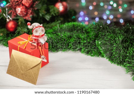 Christmas owl figurine sits on a gift in red paper next to a gold envelope which is on a white wooden table next to a green tinsel in the background a colorful garland