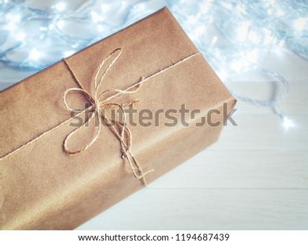 The gift is wrapped in craft paper and tied with a rough rope. On a wooden background is a luminous garland. Top view