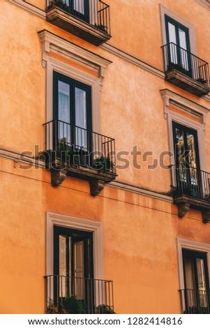 Looking up from ground level at a group of Italian apartment balcony windows and plants with an orange terracotta facade and one window lit up.