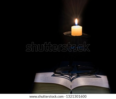 Opened book with eyeglasses, near glowing candles in candlestick, all objects are on dark background