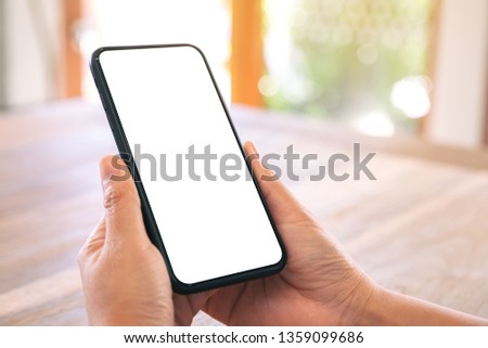 Mockup image of woman's hands holding black mobile phone with blank white screen on wooden table