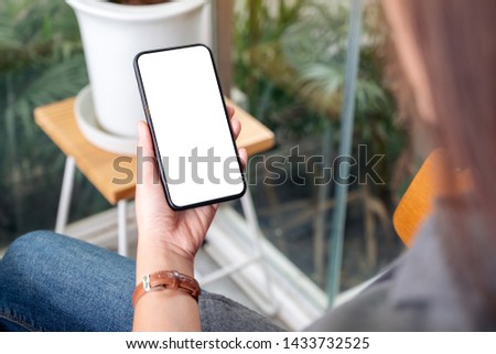 Mockup image of a woman holding and using black mobile phone with blank desktop screen in cafe