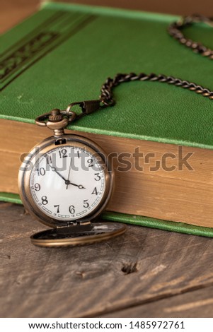 Pocket watch sitting by a book beside it. The time on the pocket watch is 3:49.