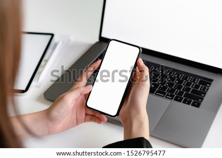 Woman holding smartphone mockup of blank screen and laptop on the table.