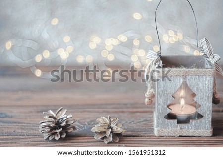 Decorative wooden candle holder with a lit candle inside it on a rustic wooden board with blurred fairy lights in the background. High key photo.