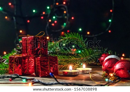 Red mini gifts on a wooden background among fir branches, lighted candles in a dark room. Beautiful, magical Christmas background with copy space.