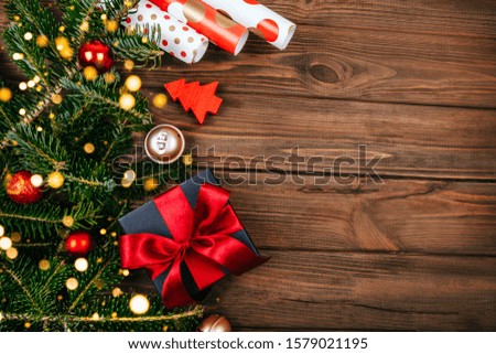 Christmas flatlay with black box and red ribbon decoration, fir tree branches, wrapping paper, tree decoration, gold bells and bokeh lightening on rustic brown wooden background.