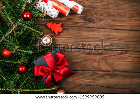 Christmas flatlay with black box and red ribbon decoration, fir tree branches, wrapping paper, tree decoration and gold bells on rustic brown wooden background.