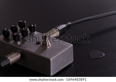 Metallic stompbox guitar effect with guitar cable and silver guitar pick on black background