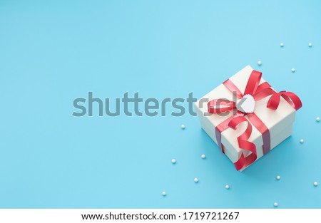 White giftbox with red ribbon and white heart on blue background with beads and copy space for text. Mother day, Valentine day, Wedding, Birthday, Christmas concept. Greeting or invitation card.