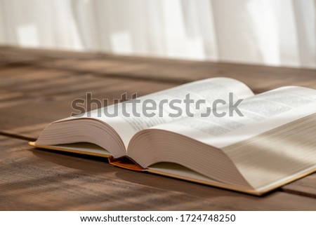 The open thick book standing on the wooden table in front of the window. Sunlight falls on the book.