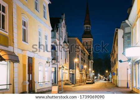 Parnu, Estonia. Night View Of Nikolai Street With Old Houses, Restaurants, Cafe, Hotels And Shops In Evening Night Illuminations. View Of Lutheran Church Of St. Elizabeth On Background