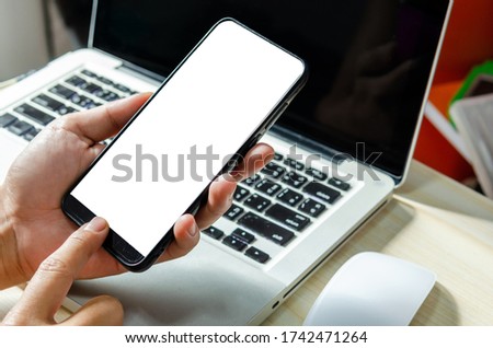 Man hand is pressing the smartphone with a blank screen for designing or advertising on the work desk and the laptop computer on the work table.