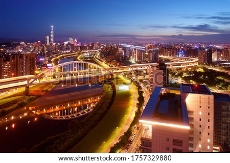 Aerial skyline of Taipei City at dusk, viewed from above a riverside park, with a highway bridge spanning Keelung River & 101 Tower standing out amid skyscrapers in Xinyi District under twilight sky
