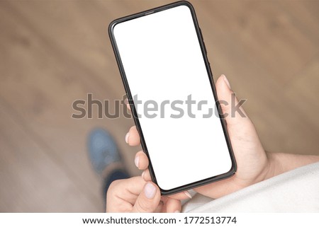 Top view mockup image of a woman holding a black mobile phone with blank white screen. Wooden floor background.