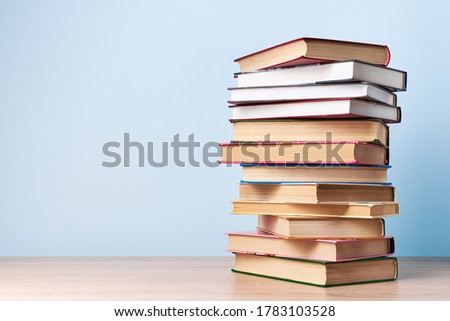 A tall, chaotic stack of books stands on a wooden table against a light blue wall, free space for text