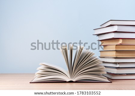 An open book and a stack of books are located on a wooden table against a light blue wall, free space for text