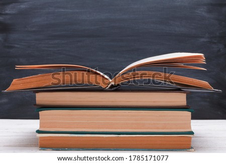 Stack of books with open book on top on white wooden desk against blackboard  background. Side view, copy space, close-up. Learning, education, knowledge, library, love reading concept