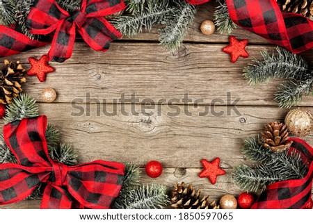 Christmas frame of ornaments, branches and buffalo plaid check ribbon. Above view on a rustic wood background.