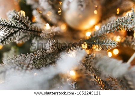 Stylish snowy christmas tree branches with golden lights close up in festive room. Modern decorated christmas tree with white balls and warm illumination, holiday wallpaper. Merry Christmas