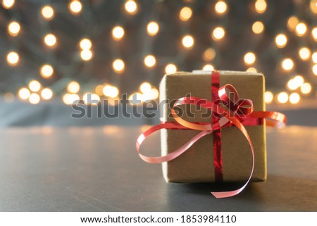 Selective focus of a bright red ribbon tied around a brown paper wrapped holiday present with bokeh Christmas lights and copy space on the left; winter