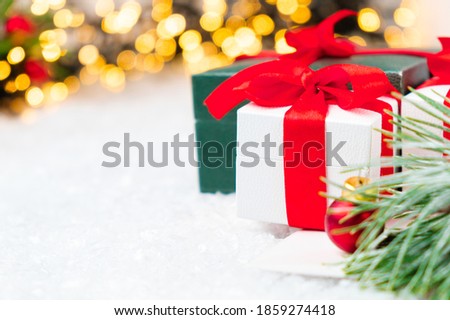 Present with red ribbon, pine or spruce branch with red balls on the snow on the background of blurred Christmas tree and lights of garland.