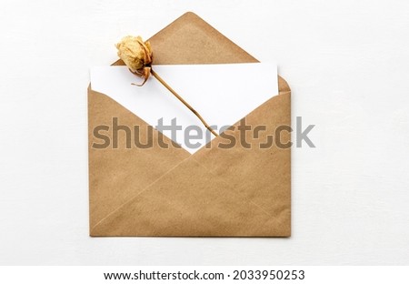 An envelope made of kraft paper, a white empty card and a dry rose on a white table. Blank or empty postcard.