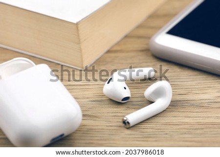 White wireless earbuds with plastic case, smarthphone and book on wooden table.
