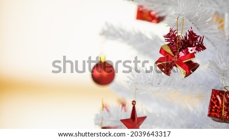 Closeup shot of glossy shiny small red bells with bow tie decorating object hang on white decoration Christmas tree branch with other star ball and gold ribbon present gift box in blurred background.