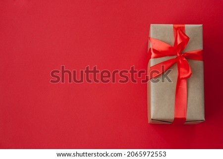 One rectangular gift box, packed in craft paper, tied with red ribbon with bow. Lies on the right on red background. View from above. Flat lay.
