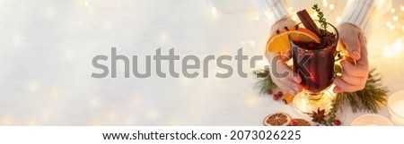 Young woman in knitted wool sweater is holding a glass with hot winter drink: mulled wine with spices, fruits. Christmas lights on background. Cozy atmosphere. Banner copy space for text