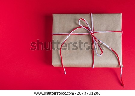 Gift parcel wrapped in craft paper, tied with a festive red and white rope, on a bright red background. Copy space view. Christmas and Happy New Year concept.