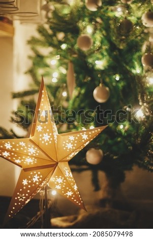Christmas star on background of christmas tree in lights in evening room. Big paper star garland glowing in festive decorated scandinavian room. Atmospheric christmas eve. Happy holidays