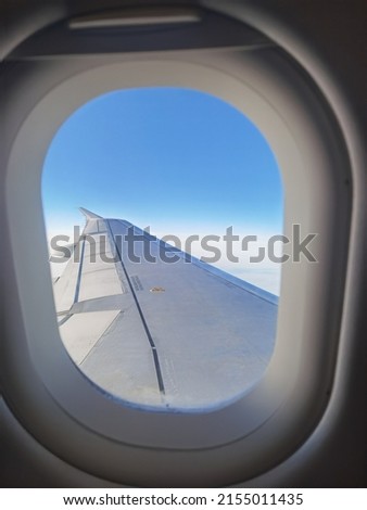 Wing of a passenger airliner flying above the clouds in a blue sky, seen through a passenger porthole