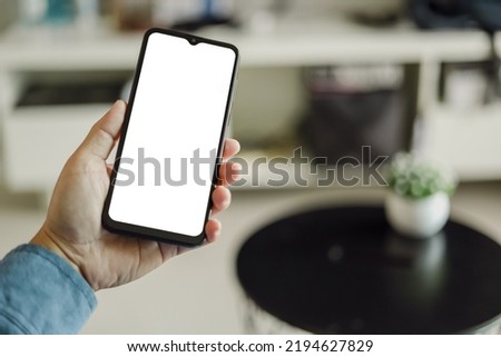 Mockup image of a man holding black mobile phone with blank white screen. In living room at home.