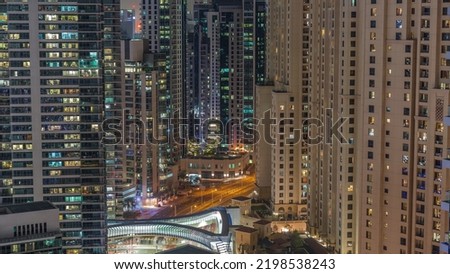 Overview to JBR and Dubai Marina skyline with glowing windows in modern high rise skyscrapers waterfront living apartments aerial night timelapse. Traffic on road intersection and footbridge