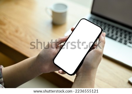 A smartphone white screen mockup is in a woman's hands over modern office desk. top view, close-up image