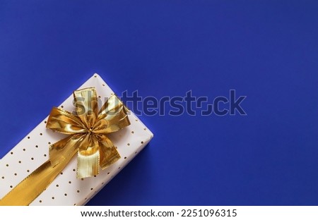 White gift box with golden ribbon and bow on a blue background. Top view. Copy space.