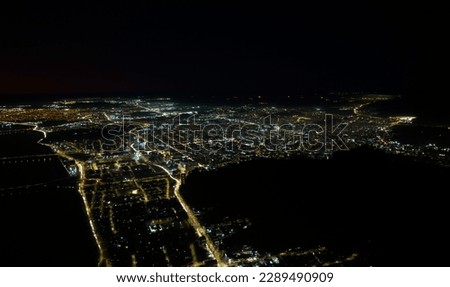 The image is an aerial view of a city illuminated at night, captured by a drone. The city is surrounded by bright lights, which spread through the streets and avenues