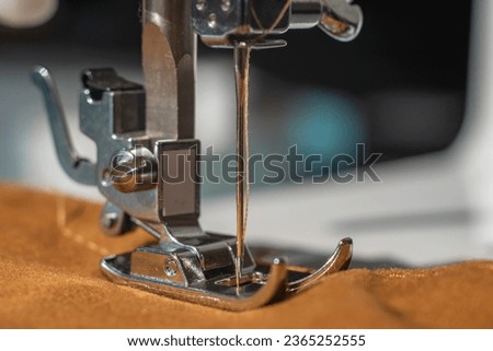 Sewing machine foot in close up.  Selective focus on the tip of the needle which is in the raised position. The needle is about to sew a brown fabric. 