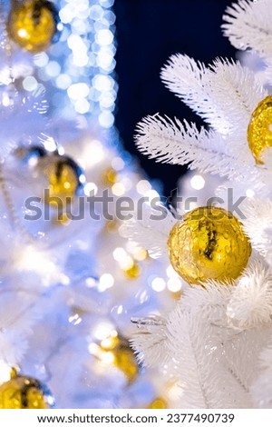 Selective focus on golden iced Christmas ball on white artificial fir branches. Defocused background with New Year decorations and lights.