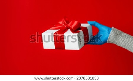 Christmas present. Festive box. Unrecognizable human hand in protective gloves holding wrapped gift isolated on red copy space background.