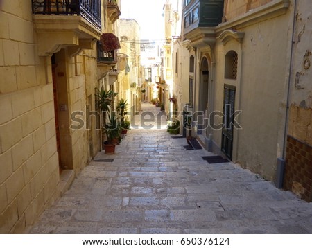 Narrow street, with plants, balcony and Maltese window box runs between limestone homes with their arched doorways and colorful doors in the Maltese city of Birgu