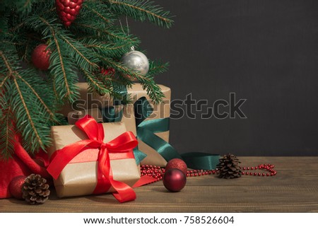 Christmas holiday background. Gifts with a red ribbon, Santa's hat and decor under a Christmas tree on a wooden board. Close up. Copy space on chalkboard.